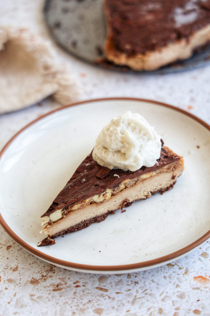 a slice of vegan snickers cheesecake on a white plate with whipped cream.